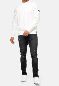 Hvid sweatshirt med præget tekst, rund hals og ribbede ærmer, parret med mørke slidte jeans og hvide sneakers.