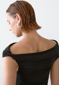 Schwarzes Off-Shoulder-Kleid mit glatter Textur und gefaltetem Design. Silberne Creolen betonen den minimalistischen Look. Kurzes, nasses Haar.