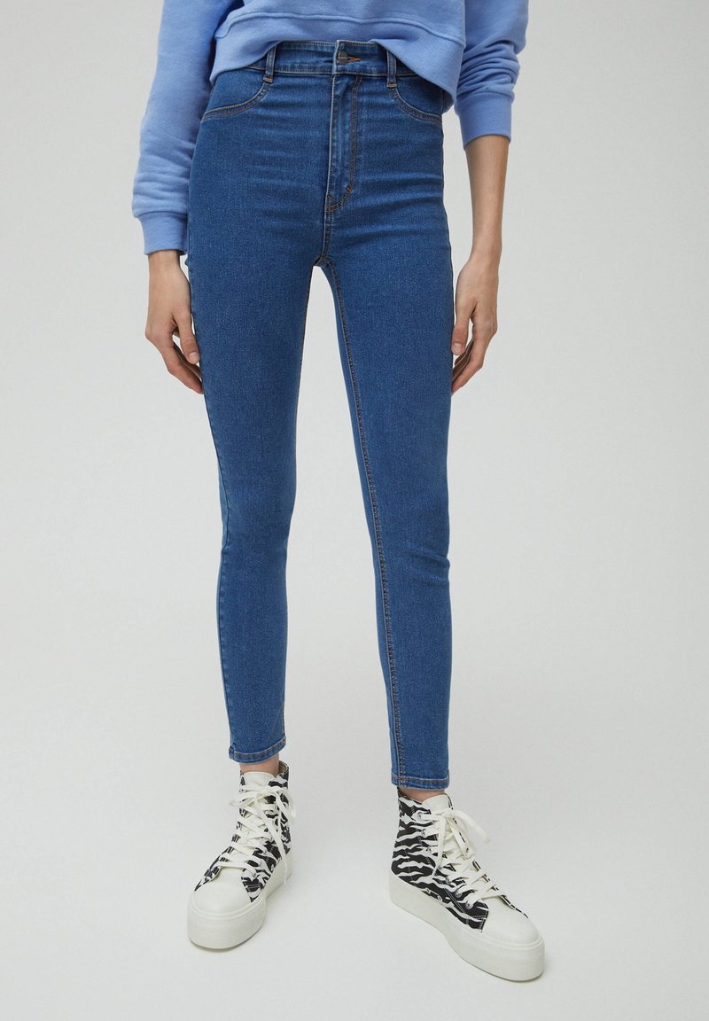 Vysoké skinny džíny v tmavě modrém denimu s hladkou texturou. Spárované s černobílými vzorovanými vysokými teniskami.