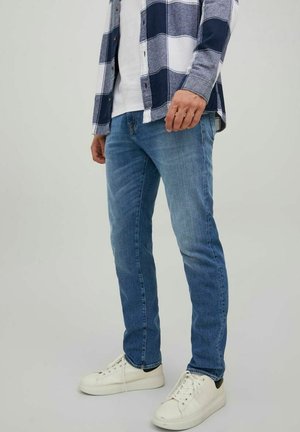 Personne portant un jean bleu, des baskets blanches, un t-shirt blanc et une chemise à carreaux bleue et blanche, debout devant un fond uni.