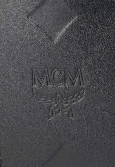 Gros plan sur du cuir noir texturé avec le logo MCM embossé et le symbole de laurier ailé.