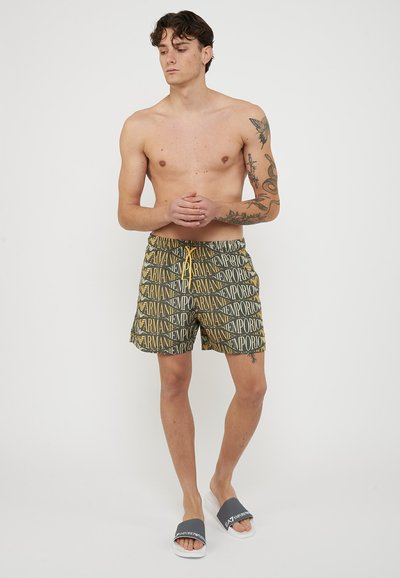 Uomo senza maglietta con tatuaggi, indossa pantaloncini da bagno a fantasia Emporio Armani e sandali EA7 neri e bianchi, in piedi con le mani congiunte.