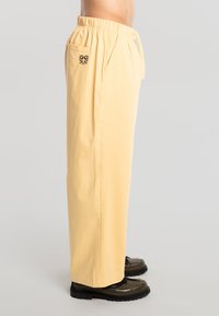 Hombre con pantalones sueltos de color amarillo pálido con cintura elástica y bordado floral negro en el bolsillo trasero, combinado con zapatos negros.