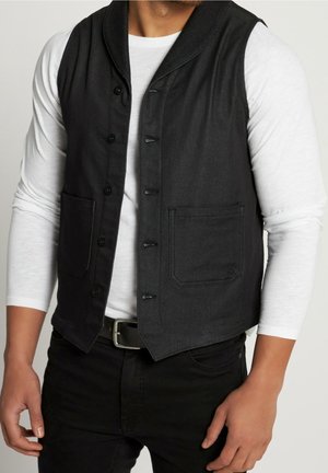 Homme portant un gilet noir boutonné sur une chemise blanche à manches longues avec un pantalon noir et une ceinture noire sur un fond neutre.