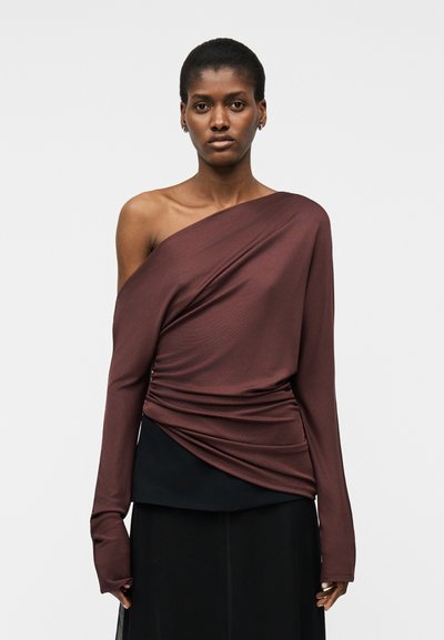 Femme portant un haut brun asymétrique à manche longue avec des détails froncés et une jupe noire sur fond blanc uni.