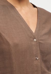 Καφέ πουκάμισο με λαιμόκοψη σε σχήμα V και δύο μπεζ κουμπιά μπροστά, φορεμένο από ένα άτομο που φαίνεται από τον λαιμό έως το μέσο του στήθους.