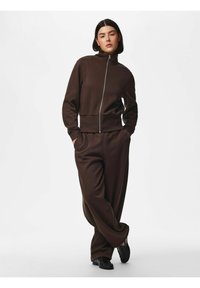 Femme aux cheveux courts et foncés, debout les mains dans les poches, portant une veste marron zippée et un pantalon large assorti avec des chaussures noires.