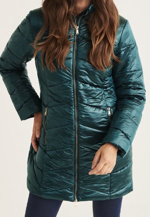 Donna che indossa una lunga giacca imbottita trapuntata color teal lucido con cerniera frontale e tasche laterali con zip, in piedi davanti a uno sfondo bianco.