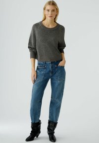 Pull en tricot gris avec des poignets larges, associé à un jean en denim bleu ample avec poches et coutures. Porté avec des bottes noires décontractées.
