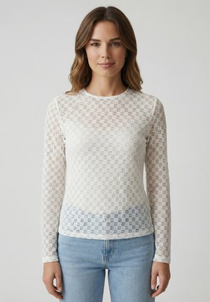 Donna con capelli castano medi, indossa una maglia bianca a maniche lunghe con motivo e jeans azzurri, su sfondo semplice.