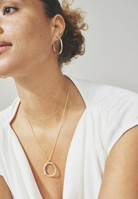 Gouden creolen en een delicate gouden ketting met een ovale hanger, gedragen over een witte blouse met een zachte textuur.