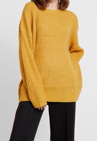 Přehnaně velký hořčicově žlutý pletený svetr s dlouhými rukávy a žebrovanou strukturou. Boční rozparky na lemu pro zvýšený komfort, kombinovaný s černými kalhotami.