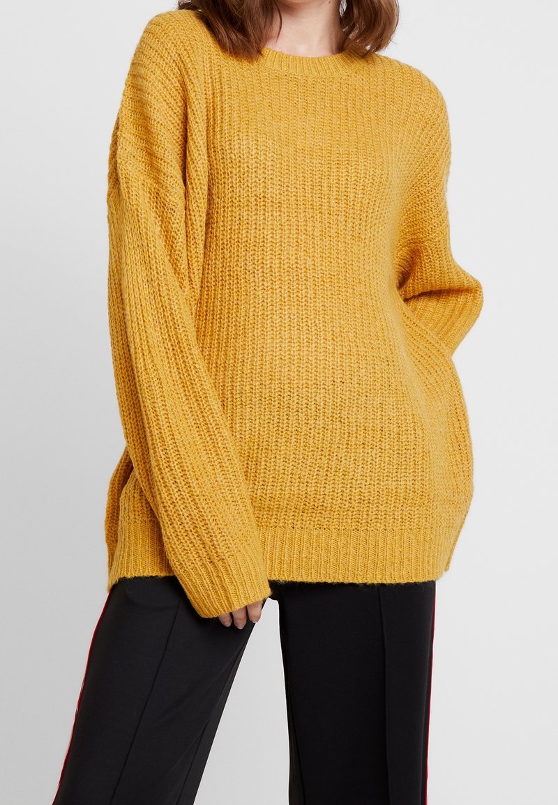 Přehnaně velký hořčicově žlutý pletený svetr s dlouhými rukávy a žebrovanou strukturou. Boční rozparky na lemu pro zvýšený komfort, kombinovaný s černými kalhotami.