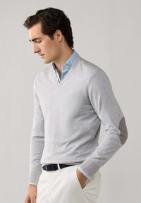 Homme aux cheveux foncés portant un pull à col en V gris clair avec des coudières, par-dessus une chemise bleue, un pantalon blanc et une ceinture noire, les mains dans les poches.