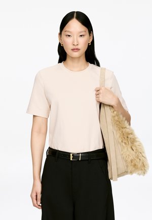 Junge Frau mit langem schwarzen Haar, die ein hellbeiges T-Shirt und schwarze Hosen trägt und eine beige Tasche mit Kunstpelzdetail auf der Schulter hält.