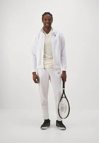 Weißer sportlicher Trainingsanzug mit Reißverschlussjacke, hellfarbenem T-Shirt und weißen Hosen. Hält einen schwarzen Tennisschläger und trägt schwarze Turnschuhe.