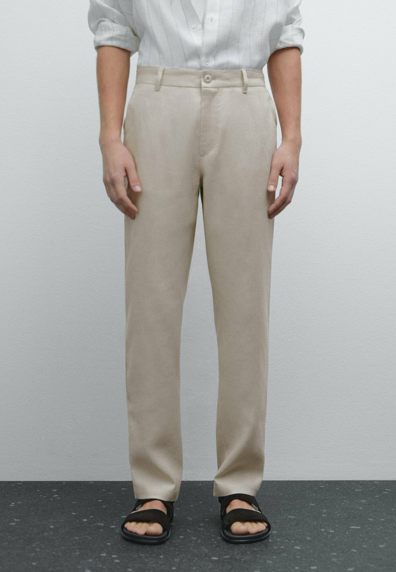 Un pantalon beige avec une texture lisse, une coupe droite, des poches avant et un léger brillant, associé à des sandales noires sur un sol texturé.