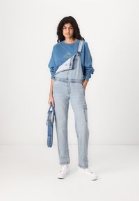Sudadera azul con gráfico de sol, tirantes de mezclilla claros con un solo tirante y bolsillo, y zapatillas blancas. Conjunto casual sobre un fondo simple.