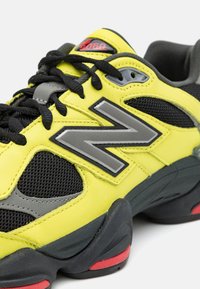 Yellow athletic shoe with gray accents, featuring a mesh and leather upper, prominent gray logo, black laces, and red sole detail.