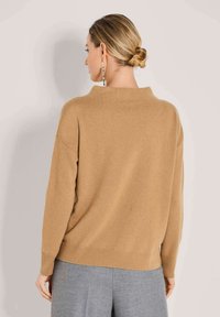 Frau mit blonden Haaren in einem tiefen Dutt, die einen camel-farbenen Pullover und graue Hosen trägt, von hinten vor einem hellen Hintergrund gezeigt.