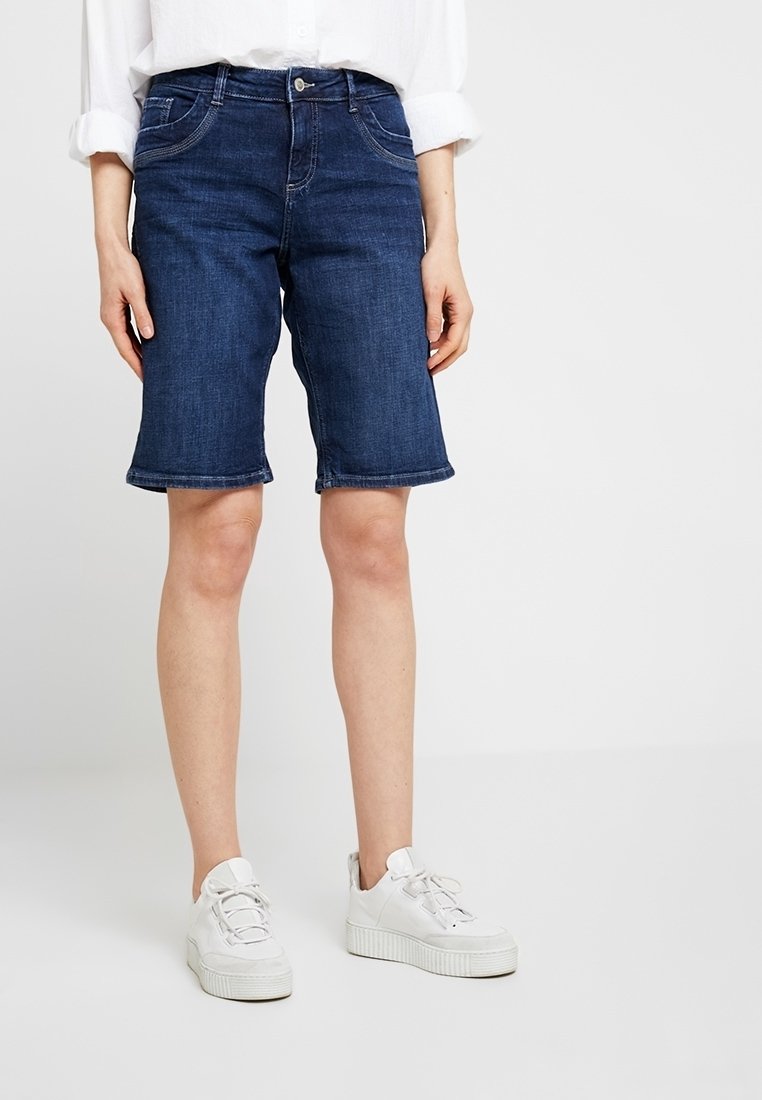 Shorts de denim en azul oscuro con corte recto y dobladillo vuelto. Combinados con zapatillas blancas. El material es algodón con un ligero estiramiento.