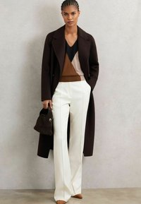 Cappotto lungo marrone, maglione a blocchi di colore in marrone, nero e beige, pantaloni bianchi su misura con cuciture laterali e una borsa scura con accentatura dorata.