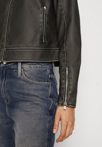 Veste en cuir noir au design court, avec des zips en argent sur les manches et les poches. Associée à un jean en denim bleu.