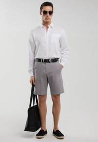 Chemise blanche en lin à manches longues, short gris, ceinture noire, sac fourre-tout noir, lunettes de soleil sombres, espadrilles ; style minimaliste et décontracté.