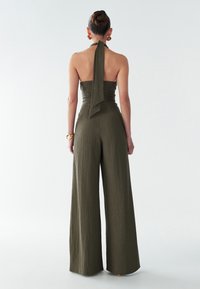 Olive green wide-leg jumpsuit with a halter neck and back tie. Textured fabric and structured fit with a loose silhouette.