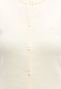 Κοντινό πλάνο σε ένα πουκάμισο μπεζ χρώματος με μικρά κουμπιά και κεντημένο λογότυπο "cK" στο αριστερό στήθος.