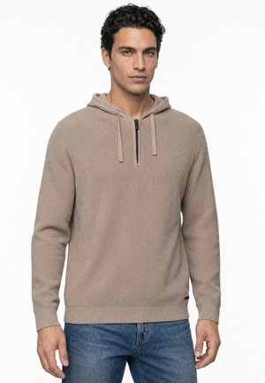 Homme aux cheveux noirs courts et bouclés, portant un sweat à capuche beige avec une fermeture éclair partielle et un jean bleu, debout devant un fond uni.