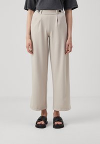 Pantaloni beige a gamba larga con pieghe davanti, dotati di due bottoni neri in vita. Abbinati a sandali neri con fasce incrociate.