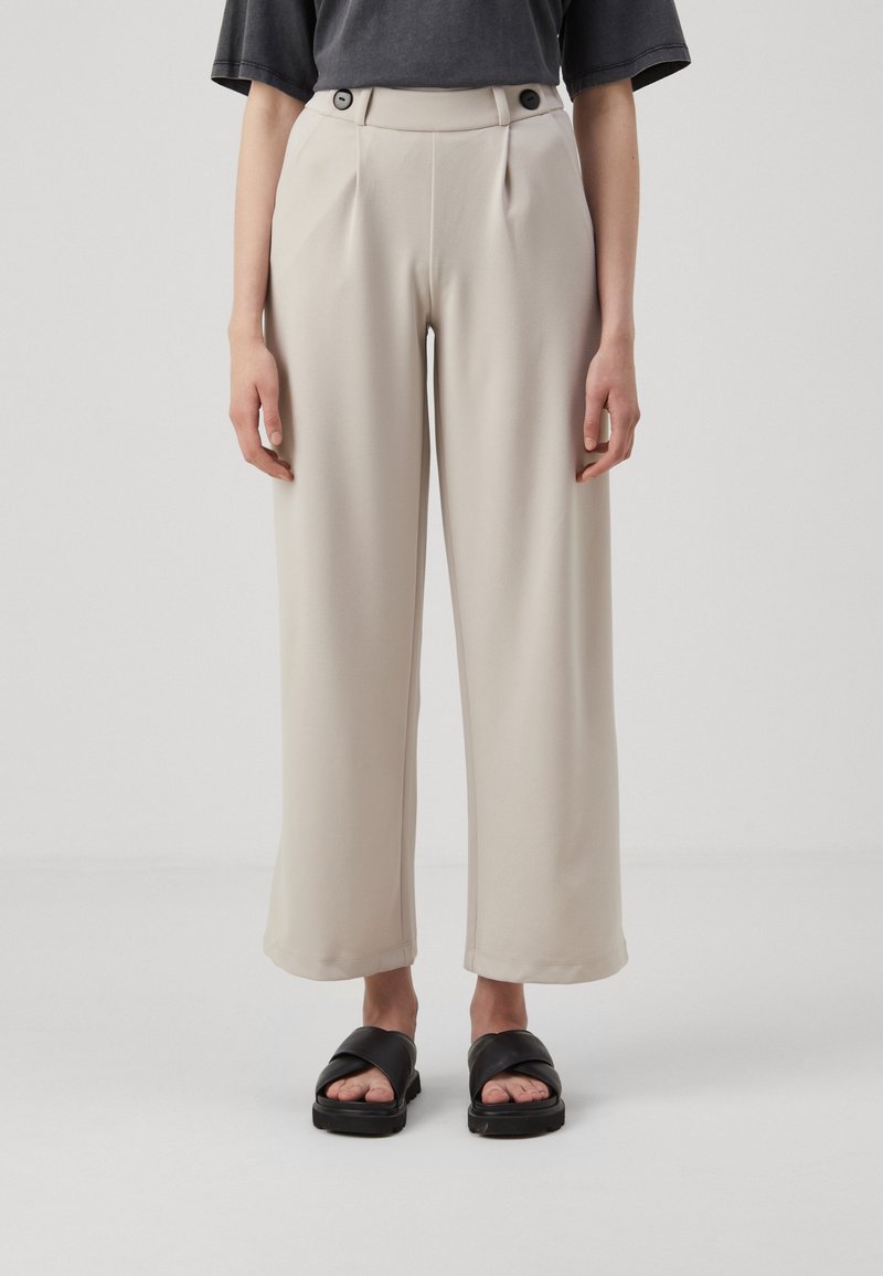 Pantaloni beige a gamba larga con pieghe davanti, dotati di due bottoni neri in vita. Abbinati a sandali neri con fasce incrociate.