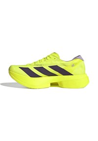 Jasnozielone buty sportowe z fioletowymi paskami, teksturowana cholewka, zaokrąglona podeszwa oraz fioletowa pętla na pięcie; charakteryzują się lekką konstrukcją i oddychającym materiałem.