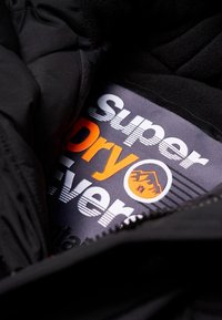 Svart jacka inuti med en grå etikett som har "Super Dry Ever" i fet vit och orange text, tillsammans med en berggrafik och randigt mönster.