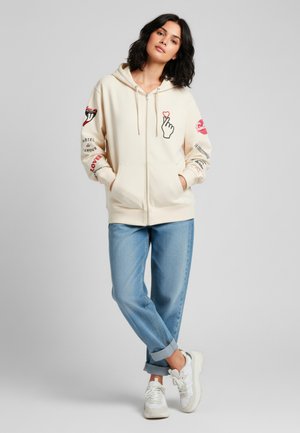 Felpa con zip color crema in misto cotone, caratterizzata da stampe grafiche sulle maniche, tasca frontale e cappuccio con cordino. Indossata con jeans rilassati e sneaker.