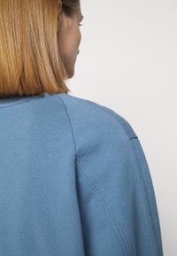 Pull en tricot bleu à texture lisse, avec des manches raglan et un col épuré, vu de dos montrant une coupe décontractée.