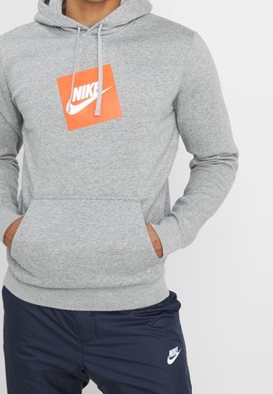 Personne portant un sweat à capuche Nike gris avec un carré logo orange et un pantalon de survêtement Nike bleu marine avec des logos blancs. Mains dans les poches du sweat à capuche.