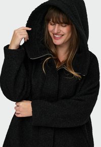 Črn plašč s kapuco, izdelan iz teksturiranega materiala, v prenapolnjenem kroju in z zadrgo. Ima širok ovratnik in prijeten, mehak videz.