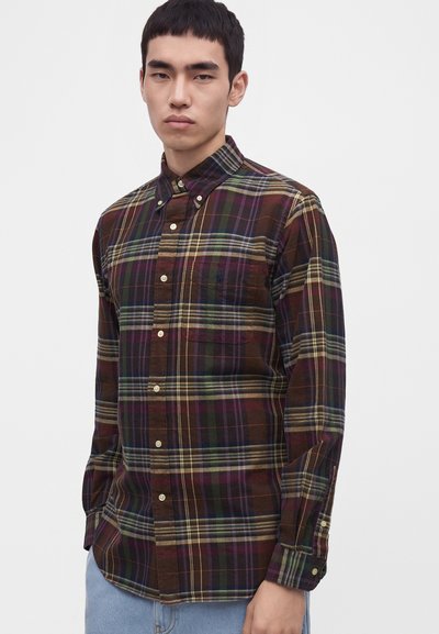 Jeune homme portant une chemise à carreaux marron, verte et violette à manches longues boutonnée avec un jean, sur un fond clair uni.