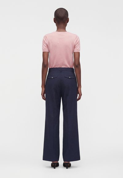 Personne debout, vue de dos, portant un haut rose à manches courtes, un pantalon large bleu marine avec poches arrière, et des chaussures noires à talons.