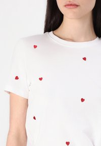 Biała bawełniana koszulka z małym czerwonym haftem serca rozrzuconym po przodzie. Krótkie rękawy i klasyczny dekolt w serek.