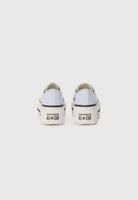 White canvas sneakers with a low profile, featuring black accents and a prominent logo on the heel. The sole is slightly elevated.