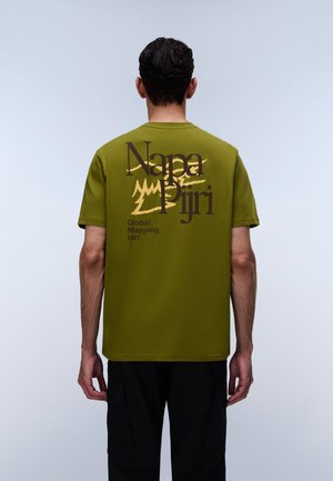 Mand iført olivengrøn T-shirt med teksten "Napa Piiri Global Mapping 1987" og grafik på bagsiden, stående mod en ensfarvet lys baggrund.