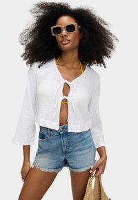 Camicetta bianca accorciata con dettaglio a nodo frontale, abbinata a pantaloncini di jeans blu. Accessoriata con occhiali da sole beige e una borsa intrecciata.