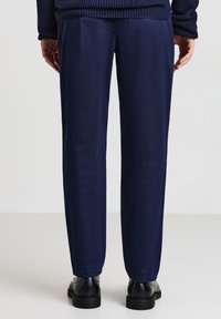 Vista posteriore di una persona che indossa pantaloni blu navy, scarpe in pelle nere e un maglione di maglia blu scuro, in piedi contro uno sfondo semplice.