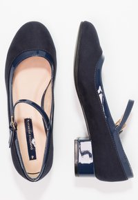 Ballerine blu navy con esterno in suede, dettagli in pelle verniciata e piccolo tacco a blocco. Presentano una cinghia con fibbia color oro.