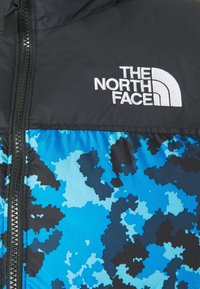 Péřová bunda s černou horní částí a modrým maskáčovým vzorem dole. Má přední zip a vyšívané logo na hrudi.