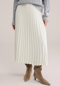 Sieviete valkā baltu plisētu midi svārku, pelēku ribotu džemperi, sudraba aproci un brūnas zamša augstpapēžu zābakus, stāvot uz neitrālas fona.