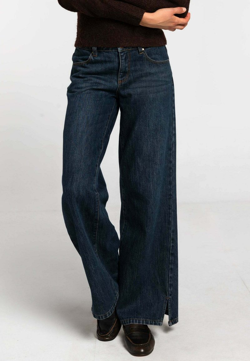 Jean en denim foncé à jambes larges avec un ourlet brut et des fentes latérales. Comprend un design classique à cinq poches et une fermeture par bouton à la taille.